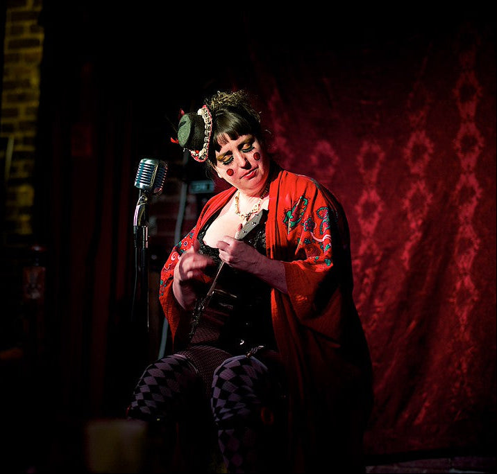 Person in a red outfit with a microphone and ukulele on a dark stage